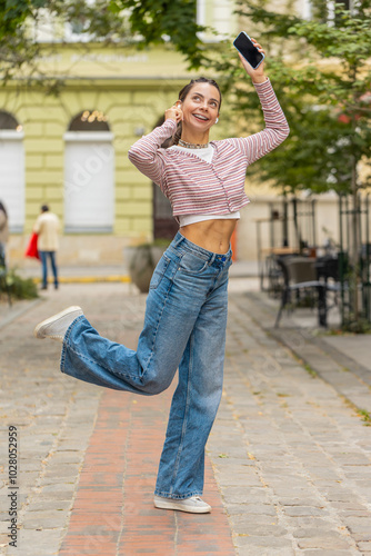 Happy relaxed overjoyed young adult girl in wireless earphones choosing, listening favorite energetic disco rock n roll music dancing outdoors. Caucasian woman walking on urban city summer street.