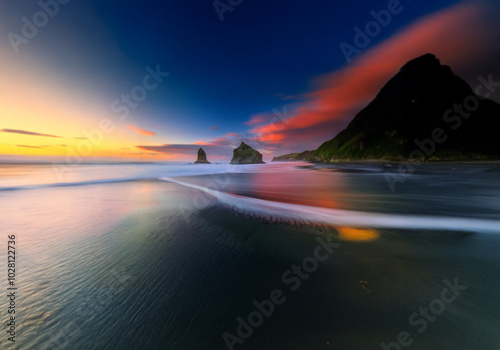 Ocean waves lap at a black sand beach, lit by a sunset sky, two rock formations jut out of the water.
