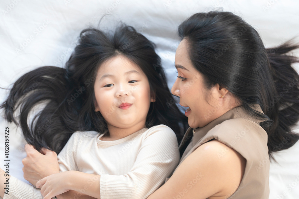 A joyful mother and daughter laughing together while lying on a bed. The playful moment showcases their strong bond and happiness in a comfortable home setting, surrounded by natural decor. A joyful mother and daughter laughing together while lying on a bed. The playful moment showcases their strong bond and happiness in a comfortable home setting, surrounded by natural decor.
