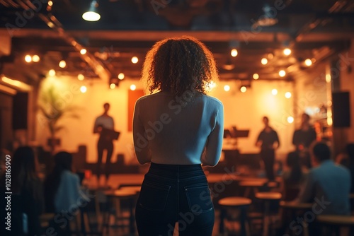 A woman stands in a dimly lit room, facing a stage with a band performing, surrounded by other audience members.