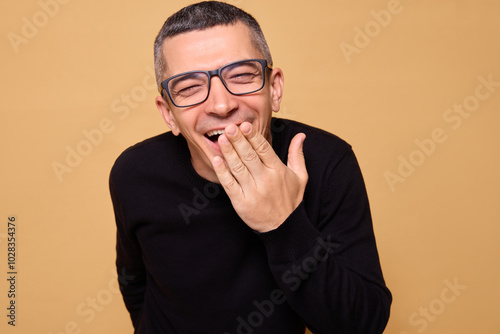 Happy man in glasses wearing jumper laughing out loud indoors covering mouth while enjoying funny joke hysterically while sharing silly anecdote isolated over beige background