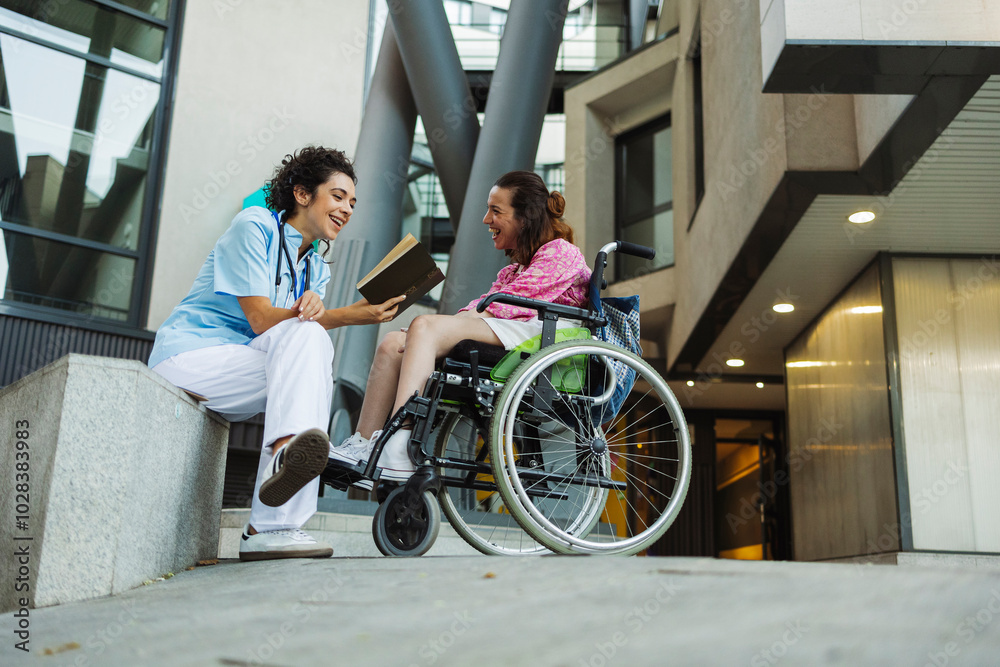 Happy assistant nurse reading book to woman sitting on wheelchair outside building Happy assistant nurse reading book to woman sitting on wheelchair outside building