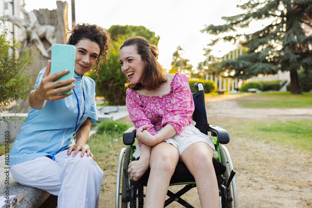 Assistant nurse in scrubs taking selfie through smart phone with woman sitting on wheelchair Assistant nurse in scrubs taking selfie through smart phone with woman sitting on wheelchair