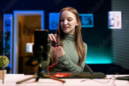 Joyous gen Z entertainer presenting wired gaming mouse from brand sponsoring video, filming with mobile phone. Upbeat tech guru promotes computer peripherals, recording with smartphone on tripod photo