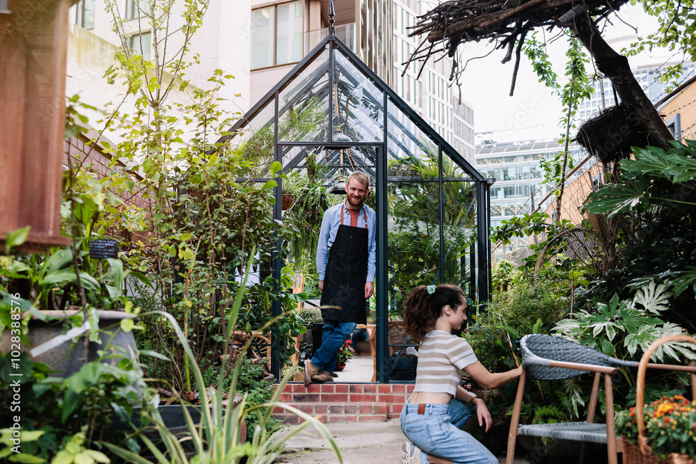 Smiling botanist standing near door and looking at girlfriend examining plants at garden centre Smiling botanist standing near door and looking at girlfriend examining plants at garden centre