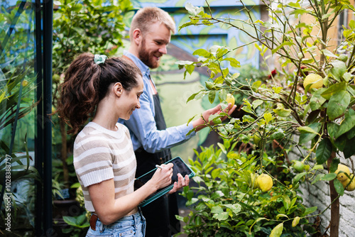 Young botanist writing on tablet PC with co-owner examining plant in greenhouse