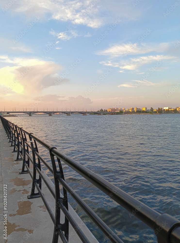 Fototapeta premium Embankment of Voronezh River is illuminated by rays of setting sun. There is road bridge inbackground. Metal barriers have been installed along embankment. Voronezh, Russia – June 17, 2024