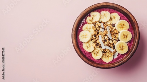 Top view of a pink smoothie bowl with sliced bananas, granola, and coconut flakes in a wooden bowl on a pink background.