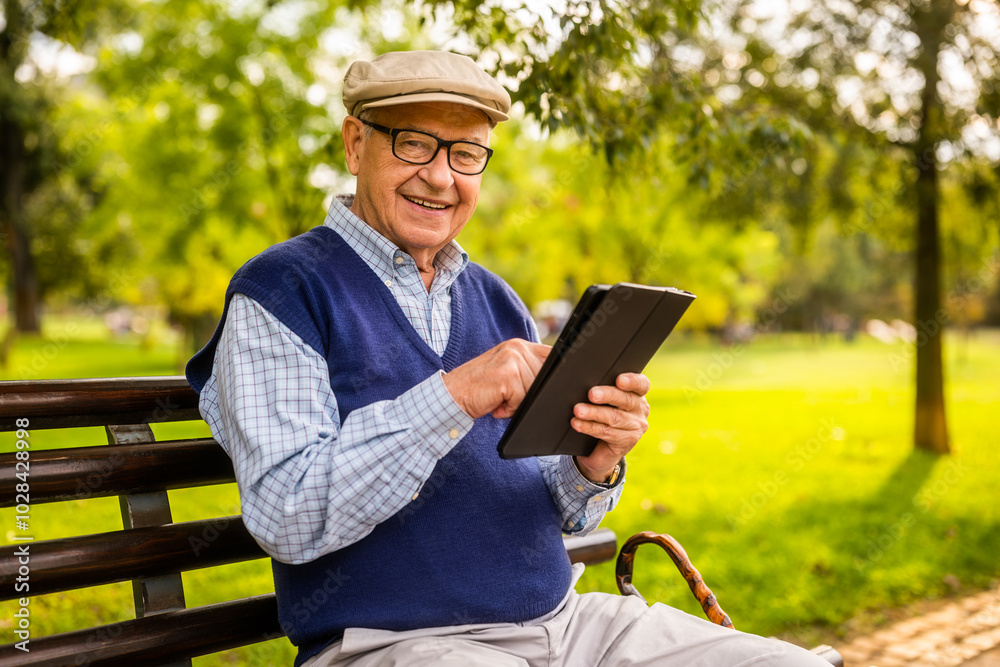 Fototapeta premium Portrait of happy senior man in park. He is sitting on bench and using digital tablet.