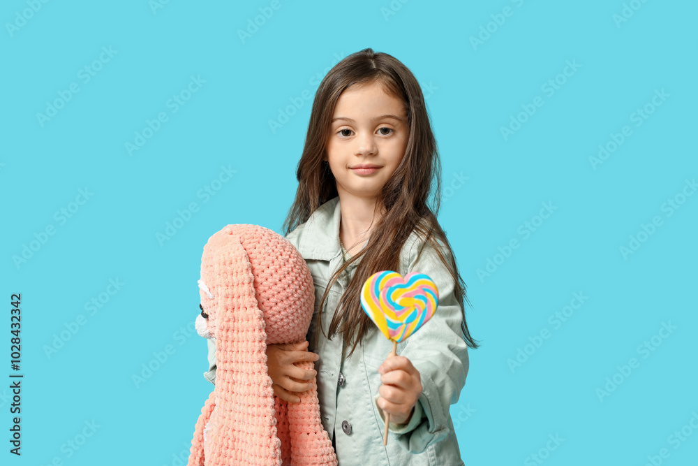 Cute little girl in stylish clothes with bunny toy and sweet lollipop on blue background