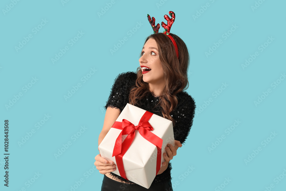 Young woman in reindeer horns with Christmas gift box on blue background
