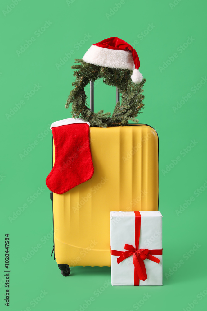 Fototapeta premium Suitcase with Christmas wreath, Santa hat, sock and gift on green background