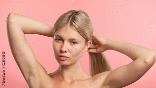 Young blonde woman posing at camera, makes ponytail and touching silky hair, isolated on pink studio background. Beauty skin care concept