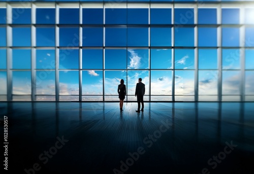 Two businesspeople stand in silhouette looking out of a large window at a cityscape.