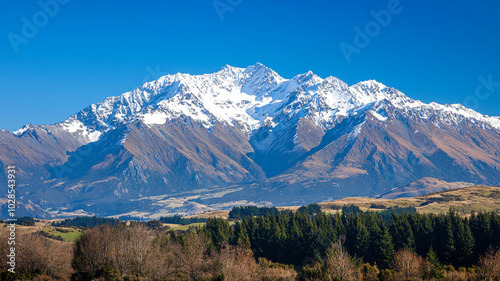 Majestic mountain range with snow capped peaks under clear blue sky, surrounded by lush greenery and rolling hills, creating breathtaking natural landscape