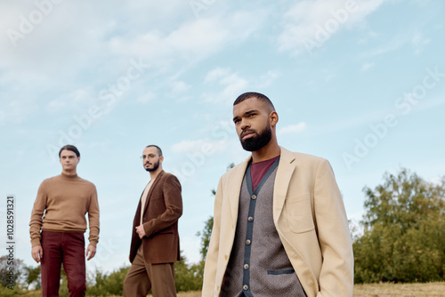 Three handsome men in fashionable autumn attire pose confidently in a tranquil field under clear skies.