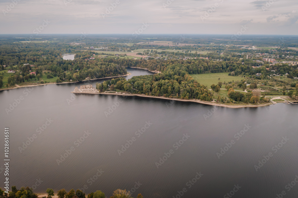 Fototapeta premium Aerial view of a calm lake surrounded by lush green forest and small settlements on a cloudy day..