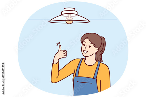 Woman electrician showing thumbs up after fixing chandelier or replacing light bulb under ceiling. Girl in overalls works as electrician efficiently performing tasks of setting up electricity