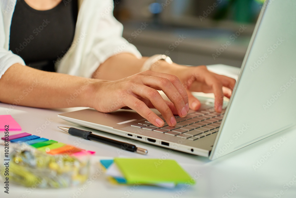 Woman working from home office typing on laptop keyboard with colorful sticky notes and stationery on desk Woman working from home office typing on laptop keyboard with colorful sticky notes and stationery on desk