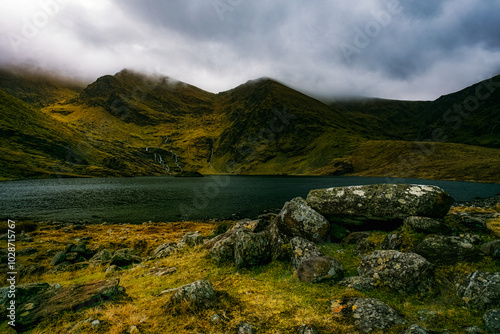 A quiet, serene lake surrounded by green hills. Large rocks litter the foreground, while a thin waterfall trickles down the mountainside. photo