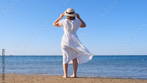 Fotografie back view on a mature woman in a straw hat and white dress walks along the blue sea coast on a sunny summer day, enjoying freedom and relaxation