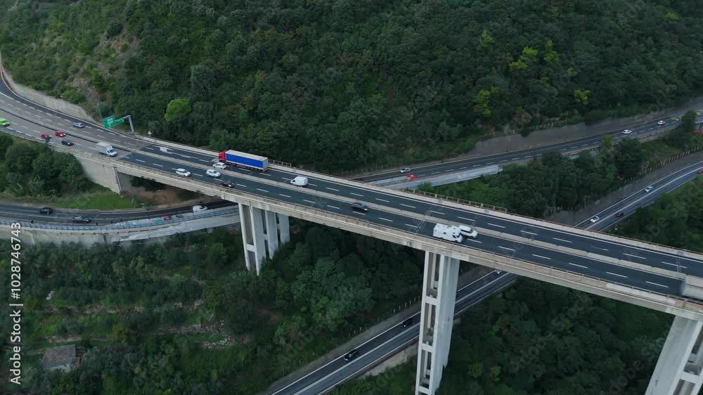 Trucks and cars crossing a large highway bridge through a green hilly landscape, aerial view Trucks and cars crossing a large highway bridge through a green hilly landscape, aerial view