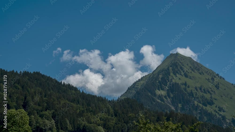 Das Zeitraffer-Video zeigt eine malerische Berglandschaft an einem klaren, sonnigen Tag. Ein grüner, bewaldeter Hügel im Vordergrund und ein steiler Berggipfel, der sich in den blauen Himmel erhebt.