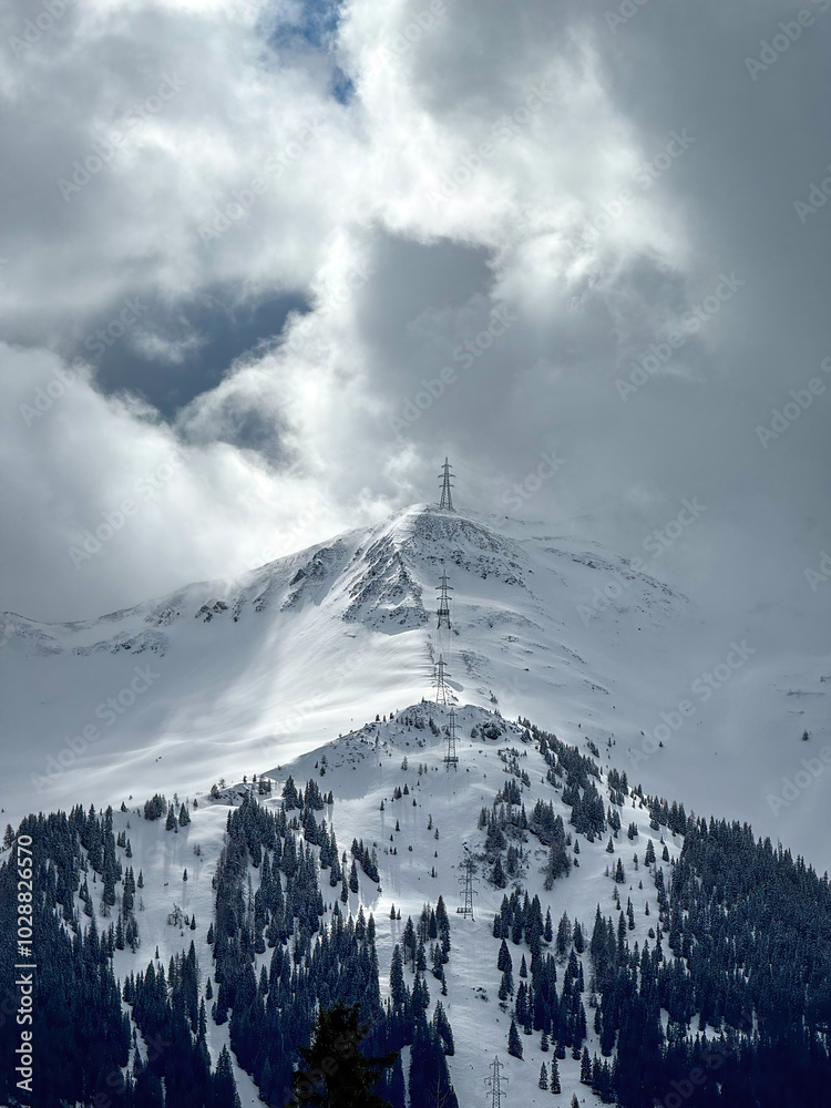 Electric power line runnnig up a snow covered mountain with snow line at the bottom in the ski region of Saalbach-Hinterglemm, Austria against dramatic cloudy sky