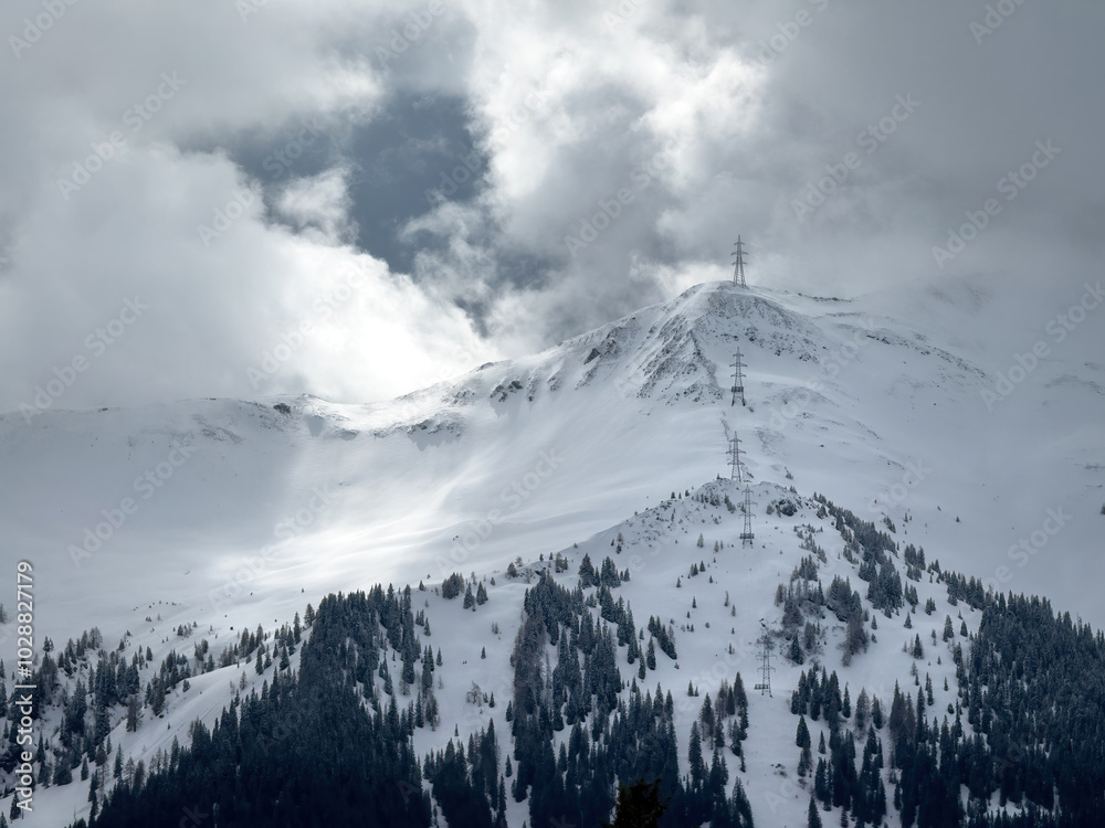 Electric power line runnnig up a snow covered mountain with snow line at the bottom in the ski region of Saalbach-Hinterglemm, Austria against dramatic cloudy sky