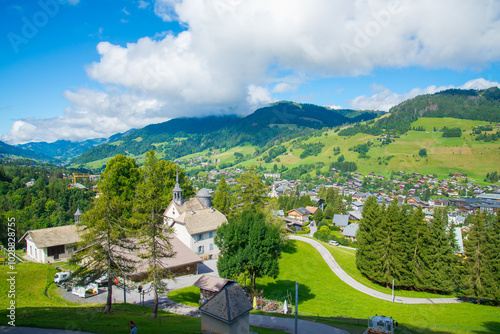 Aerial view of the village of Megeve, France in its green valley, with green hills and trees. Blue sky with white clouds on the background.