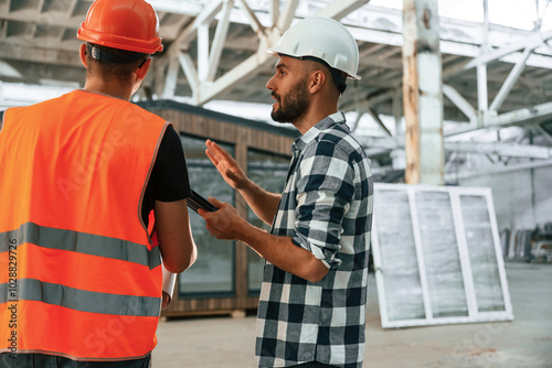 Builder is talking with manager or architect. Two workers are in the industrial wooden warehouse