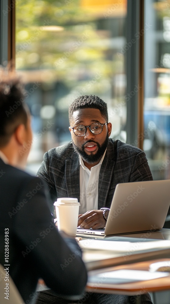 Focused professional in glasses and plaid blazer engages in serious discussion with colleague at cafe table, laptop and coffee cup nearby, city backdrop visible through window. Focused professional in glasses and plaid blazer engages in serious discussion with colleague at cafe table, laptop and coffee cup nearby, city backdrop visible through window.