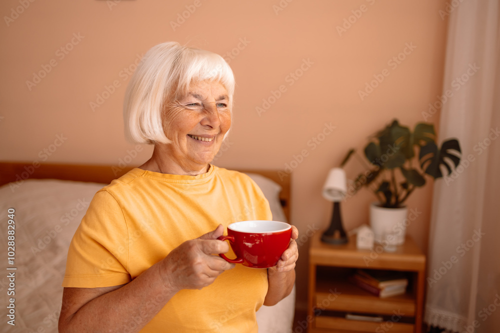 Happy Caucasian blonde senior woman sitting on a white bed with a cup in her hand. Female relaxing and drinking cup of hot coffee or tea in the bedroom.