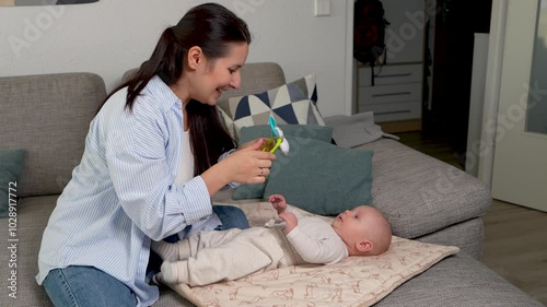 Mother holds rattle in hands. Newborn baby lying on couch and looking at toy with curiosity. Development of baby, maternal care.