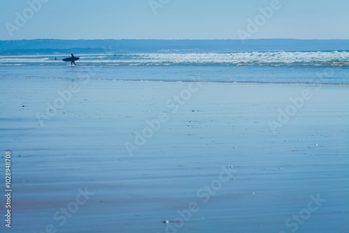 This photograph captures a peaceful moment on a wide, empty beach with a lone surfer walking along the shoreline.