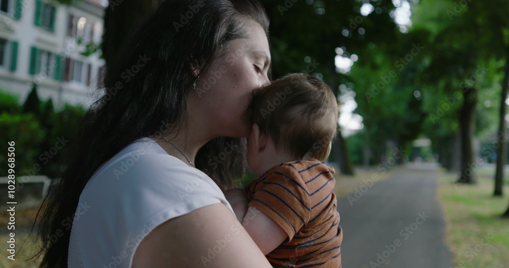 Fototapeta premium Mother kissing her baby’s head while holding him close, standing on a tree-lined path, expressing tenderness and love, with a peaceful urban setting