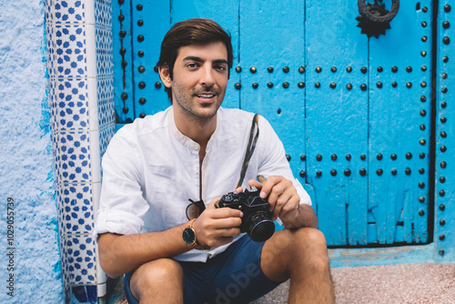 Portrait of happy hipster guy dressed in stylish apparel enjoying free time for photography hobby and discovering country with berber culture, positive man smiling at camera testing retro technology #1029055739