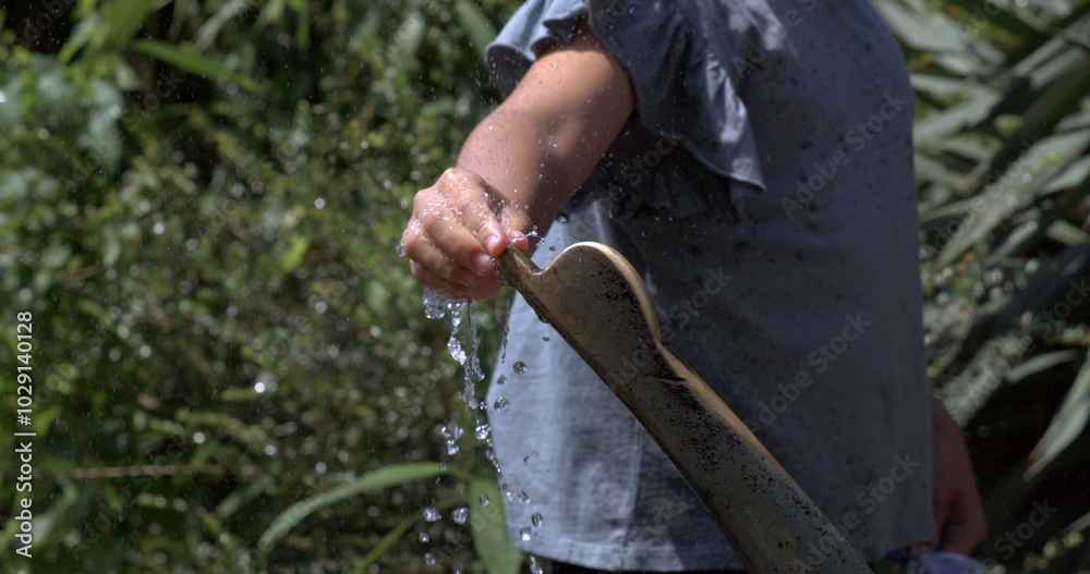 Obraz premium A child’s hand interacting with a public fountain, slow-motion capture of water droplets sparkling in the sunlight, against a lush garden backdrop