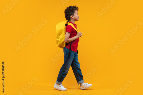 Full length studio portrait of a cute casually dressed happy smiling confident African American schoolboy walking with his bright colored backpack over orange yellow background