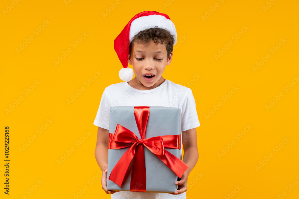 Cute little African American boy in basic white t-shirt and Santa Claus hat looking at the Christmas present box in his hands with surprised face expression, isolated on yellow background Cute little African American boy in basic white t-shirt and Santa Claus hat looking at the Christmas present box in his hands with surprised face expression, isolated on yellow background