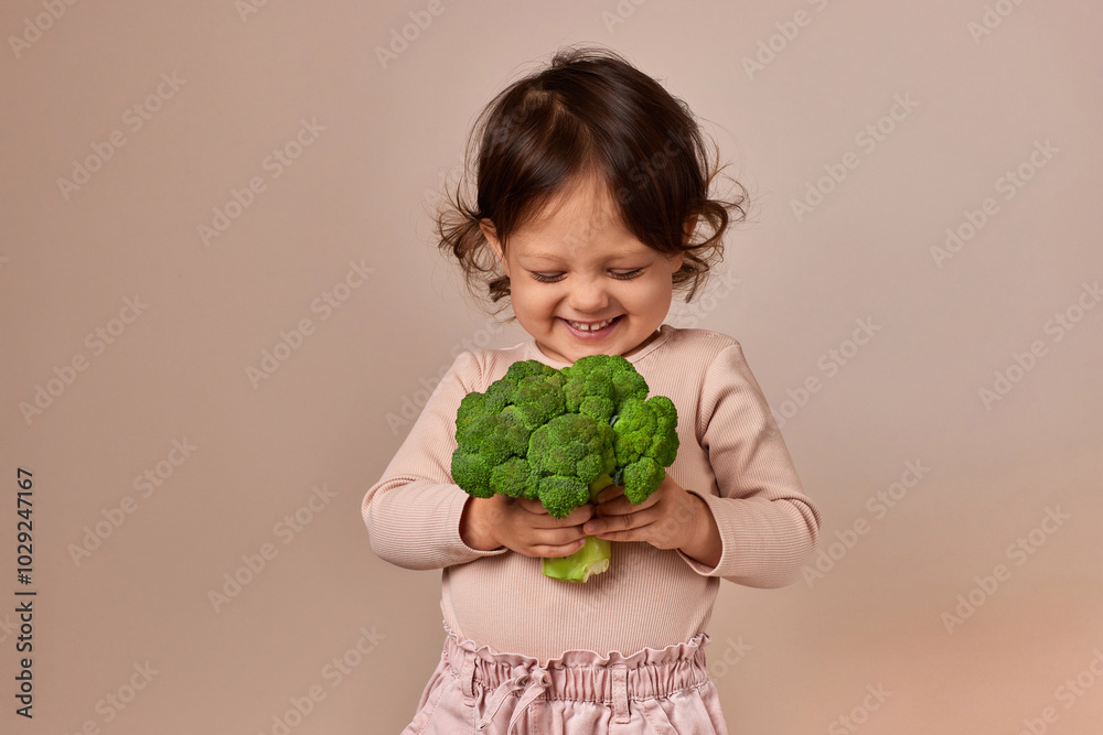 smiling cute child girl holding fresh green raw broccoli on beige background. healthy baby food. kid loves vegetables smiling cute child girl holding fresh green raw broccoli on beige background. healthy baby food. kid loves vegetables