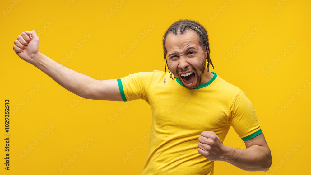 Euphoric Brazilian young footballer or soccer fan man in yellow green t-shirt cheering up making wiiners gesture clenching fists and screaming yes on yellow background Euphoric Brazilian young footballer or soccer fan man in yellow green t-shirt cheering up making wiiners gesture clenching fists and screaming yes on yellow background