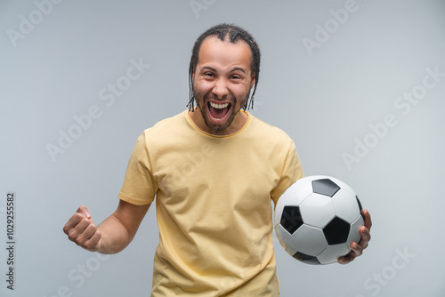 Studio shot of euphoric football fan man celebrating victory after betting at bookmaker's website, cheering, screaming yes and making winner's gesture clenching his fist while holding ball in hand