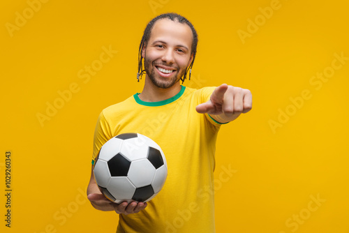 Portrait of young happy Brazilian football player or supporter posing in studio holding soocer ball and pointing with index finger directly at the camera, isolated over yellow background photo