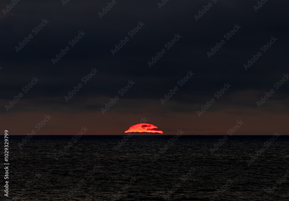 The sun is slowly setting over the vast ocean under a cloudy sky
