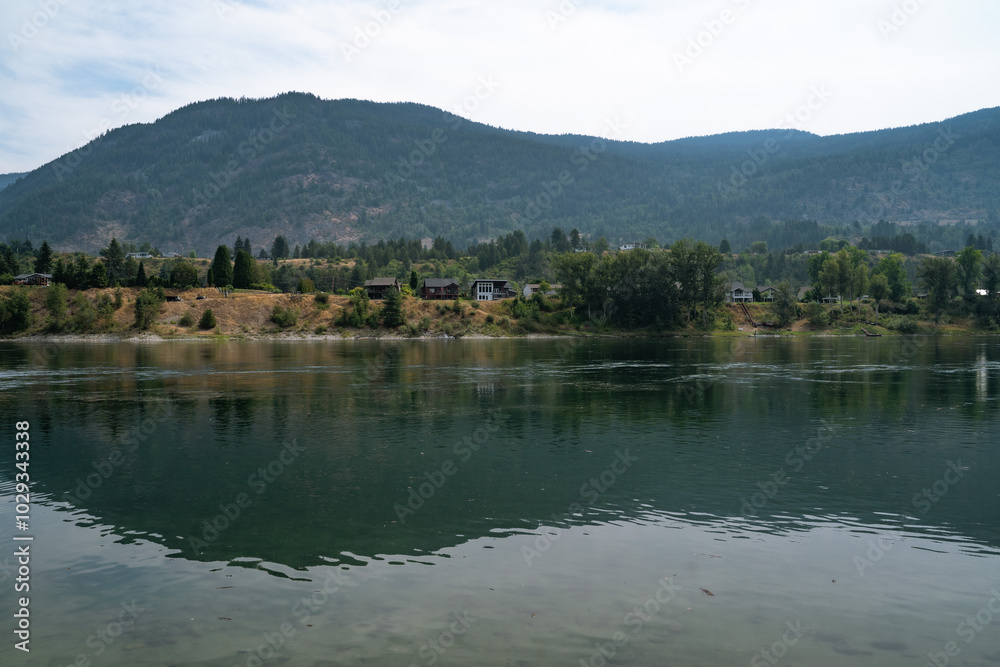 Fototapeta premium Serene river landscape with reflections of mountains and trees in the soft light of a summer afternoon near a quiet residential area