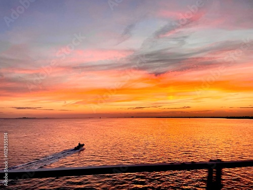 A view from the driving bridge over the calm water as the sun sets and meets the sea.  Bold, vivid colors reflect on pink orange and red water as a boat heads into the sunset at dusk, causing a ripple photo