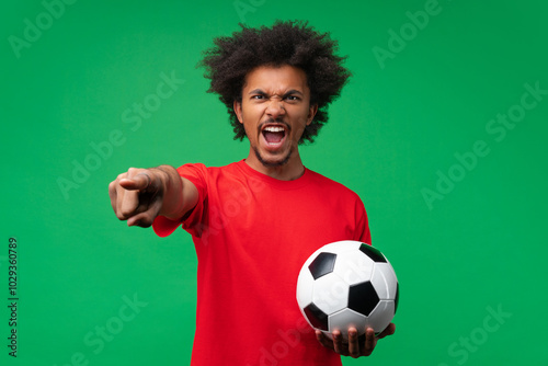 Young angry screaming african american fan man cheering up supporting football sport team holding soccer ball and pointing index finger  to the camera isolated over green background photo