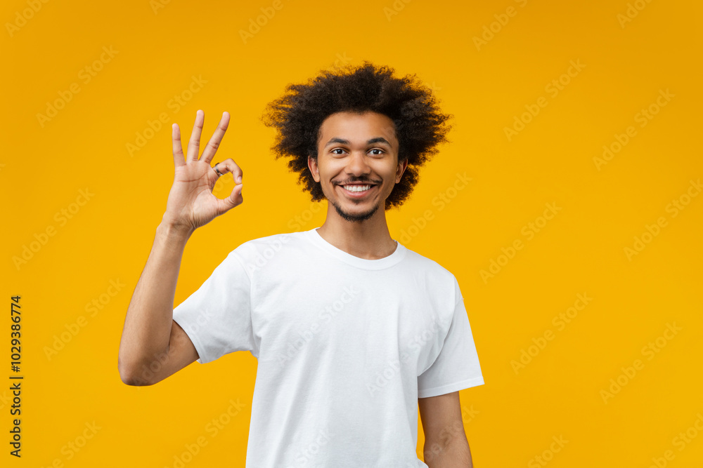 Studio shot of young happy smiling African American man in basic white t-shirt posing over bright colored orange yellow background showing ok sign gesture Studio shot of young happy smiling African American man in basic white t-shirt posing over bright colored orange yellow background showing ok sign gesture