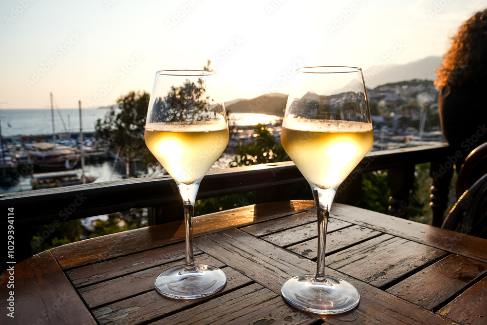 Obraz premium Drinking white wine in the bar or marine by the Mediterranean Sea at golden hour or sunset. Two glasses of white wine on the table